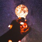 hand holding lit lightbulb. galaxy inbackground
